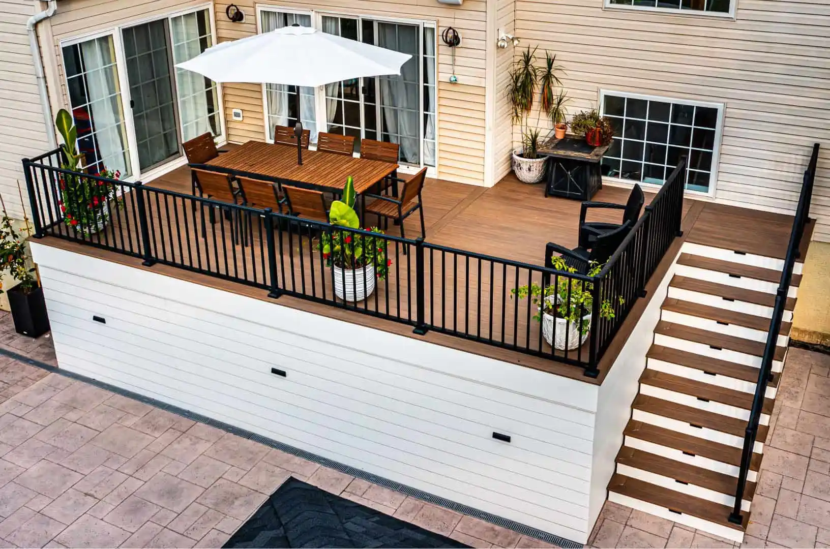 A raised wooden deck features a dining table, chairs, and a large white umbrella. Planters with greenery adorn the corners, while black metal railing surrounds the space. Downstairs leads to a paved area, showcasing expert hardscaping services in Delaware County, PA by the beige house with large windows.