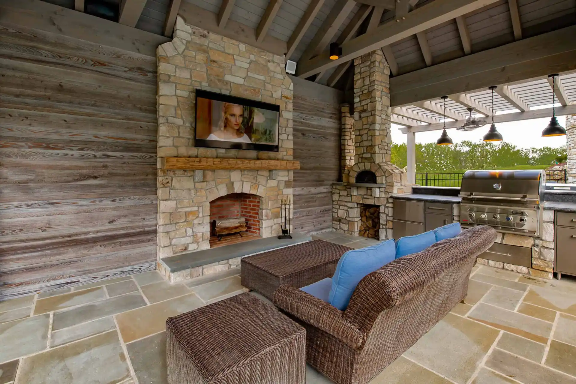 Outdoor living in Delaware County, PA, shines with a patio featuring a stone fireplace and flat-screen TV. A wicker sofa with blue cushions offers comfort facing the TV. An outdoor kitchen, complete with grill and countertops, graces the right side under a charming wooden pergola.