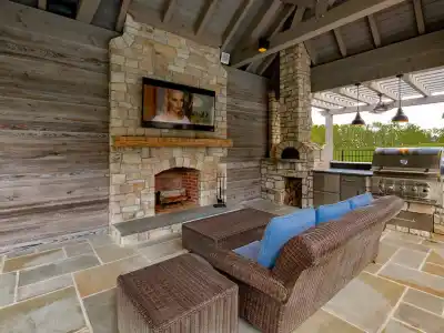 Experience outdoor living in Delaware County, PA, with a stunning patio featuring a stone fireplace and flat-screen TV. Relax on the wicker sofa with blue cushions while enjoying the stainless steel grill station under a pergola adorned with hanging lights. Perfectly designed for comfort and style!.