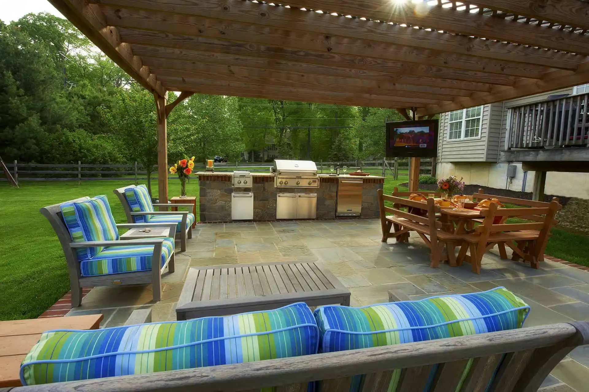 An outdoor patio with striped blue and green cushioned chairs, a wooden table and benches, a built-in grill station, and a pergola overhead, set in a lush green backyard.