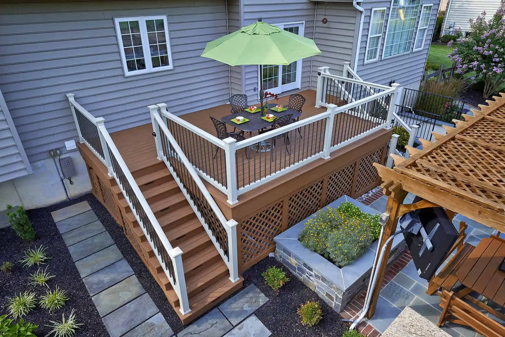 A raised wooden deck attached to a house features a dining table with four chairs and a green umbrella. Stairs lead down to a landscaped yard with stone paths, plants, and a wooden pergola.