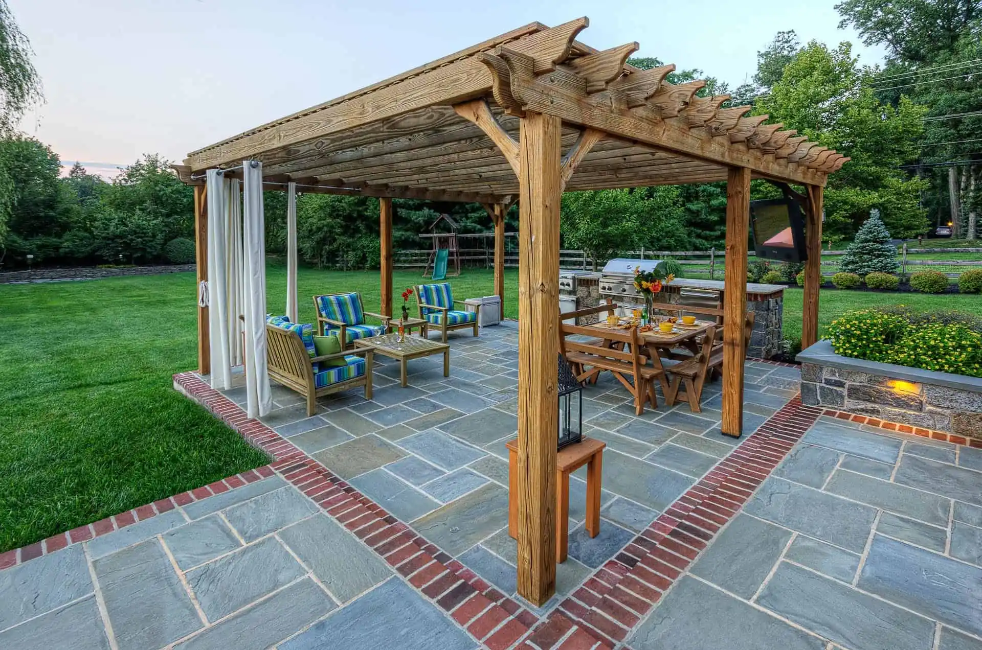 A wooden pergola covers a patio with outdoor seating and a dining table set for a meal. The stone patio is bordered by red bricks and surrounded by a lush green lawn and trees in the background.
