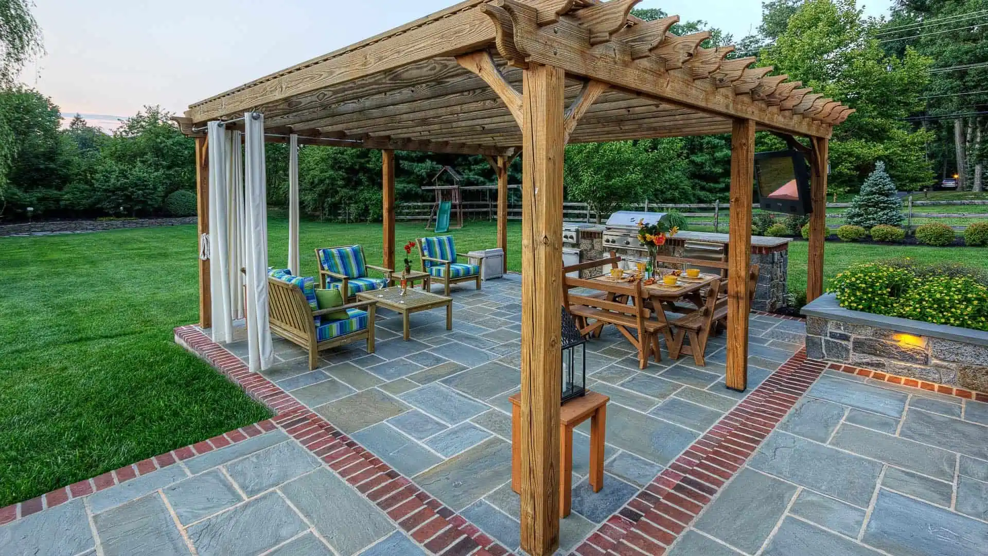 A wooden pergola on a patio with outdoor furniture, including a dining table and cushioned chairs. The area is adorned with potted plants and surrounded by a garden and lawn, crafted through expert landscaping services in Delaware County, PA. The setting is inviting and tranquil.