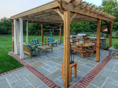 A wooden pergola on a patio with outdoor furniture, including a dining table and cushioned chairs. The area is adorned with potted plants and surrounded by a garden and lawn, crafted through expert landscaping services in Delaware County, PA. The setting is inviting and tranquil.