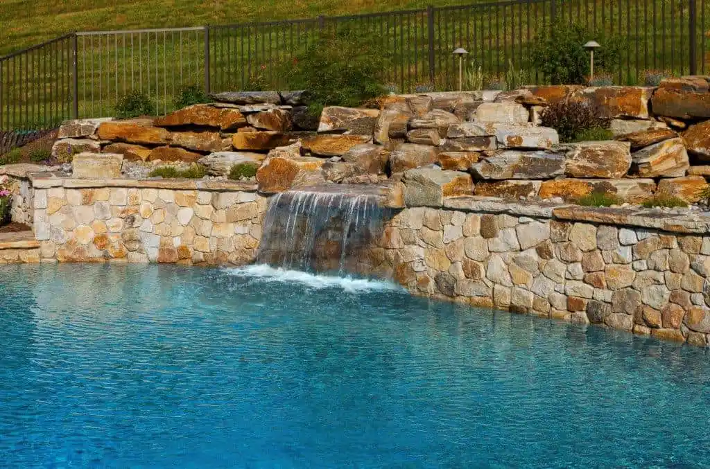 A serene swimming pool with clear blue water features a rock waterfall, beautifully showcasing the artistry of outdoor living in Delaware County, PA. The cascading waters flow over a stone wall surrounded by layered rocks, with a grassy hill and metal fence completing the picturesque scene.