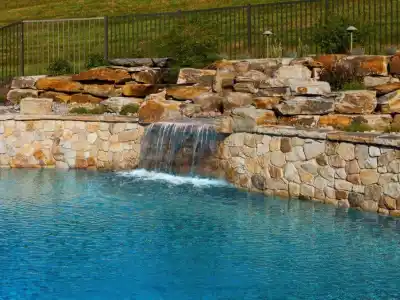 A serene swimming pool with clear blue water features a rock waterfall, beautifully showcasing the artistry of outdoor living in Delaware County, PA. The cascading waters flow over a stone wall surrounded by layered rocks, with a grassy hill and metal fence completing the picturesque scene.