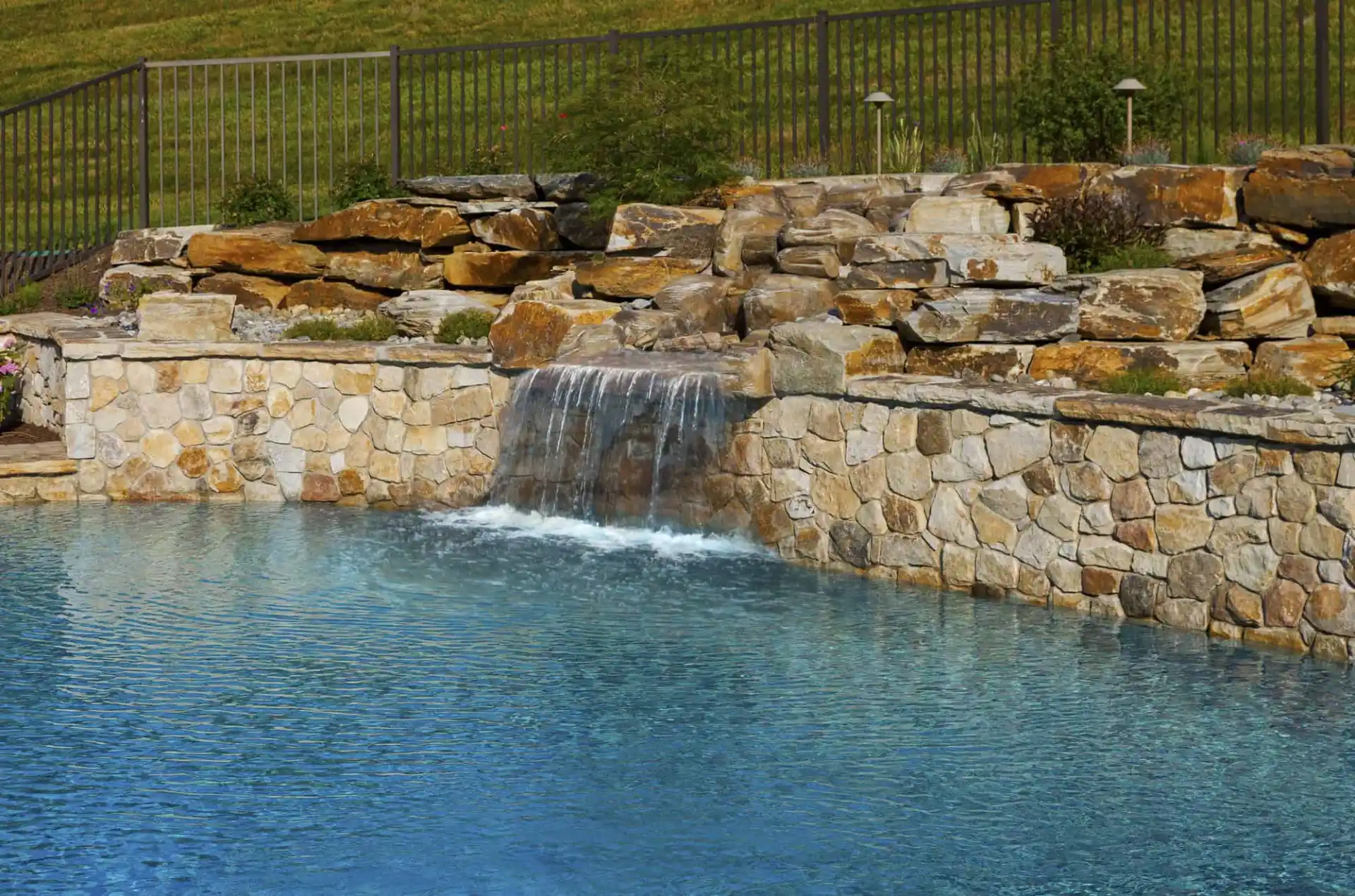 A backyard pool with clear blue water features a stone wall and a small waterfall cascading from stacked rocks, with green grass and a metal fence in the background.