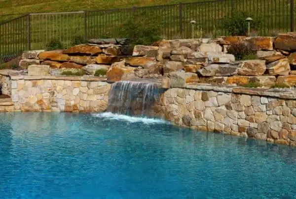 A serene swimming pool with clear blue water and a cascading waterfall feature made of stacked stones, enhanced by expert hardscaping services. The pool is surrounded by a rock wall, with a grassy hillside and metal fence visible in the background, epitomizing outdoor living in Delaware County, PA.