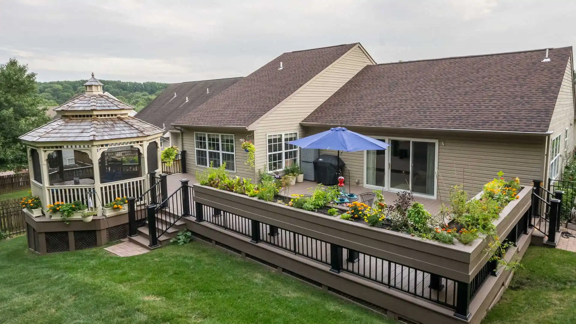 A backyard in Delaware County, PA, showcases a large wooden deck adorned with planters full of vibrant flowers and plants. A blue umbrella offers shade as a child plays nearby, with a charming gazebo to the left. The beige house boasts large windows, highlighting expert landscaping services.