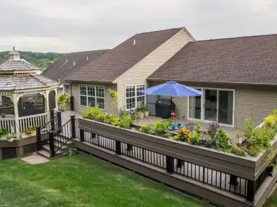 A backyard in Delaware County, PA, showcases a large wooden deck adorned with planters full of vibrant flowers and plants. A blue umbrella offers shade as a child plays nearby, with a charming gazebo to the left. The beige house boasts large windows, highlighting expert landscaping services.