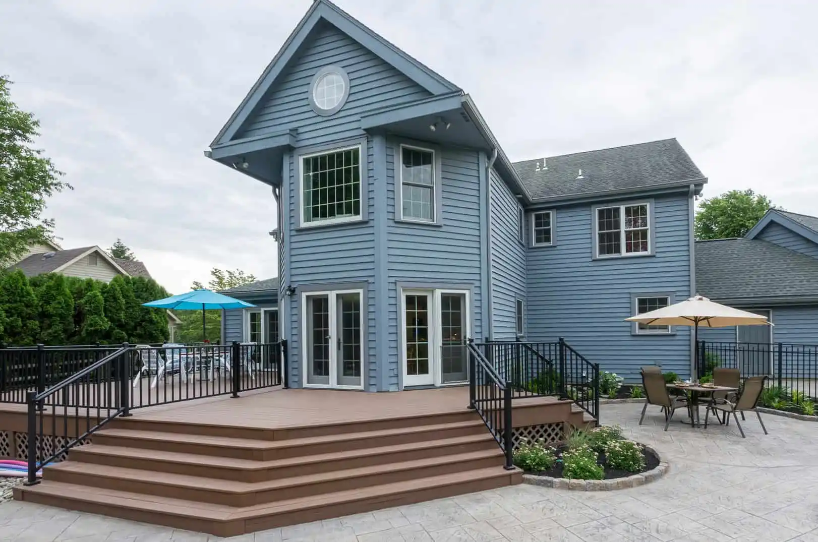 A charming two-story blue house boasting large windows and a wooden deck with stairs, black railings, and an outdoor dining set with an umbrella. Surrounded by lush greenery and a stone patio, it exemplifies exceptional hardscaping services in Delaware County, PA.