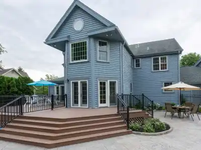 A charming two-story blue house boasting large windows and a wooden deck with stairs, black railings, and an outdoor dining set with an umbrella. Surrounded by lush greenery and a stone patio, it exemplifies exceptional hardscaping services in Delaware County, PA.