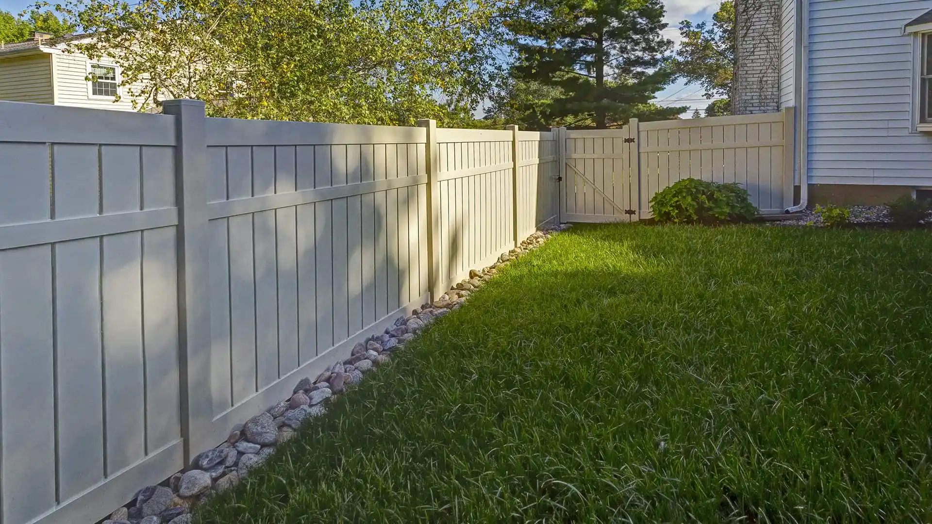 A backyard with a white wooden fence enclosing a grass lawn. Rocks line the base of the fence, and a gate is visible in the distance. Sunlight casts shadows on the lawn and fence, perfect for enjoying outdoor living in Delaware County, PA, with trees and parts of houses in the background.