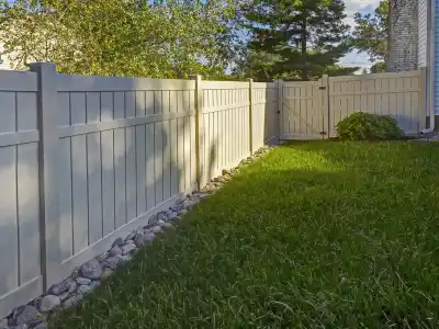A backyard with a white wooden fence enclosing a grass lawn. Rocks line the base of the fence, and a gate is visible in the distance. Sunlight casts shadows on the lawn and fence, perfect for enjoying outdoor living in Delaware County, PA, with trees and parts of houses in the background.