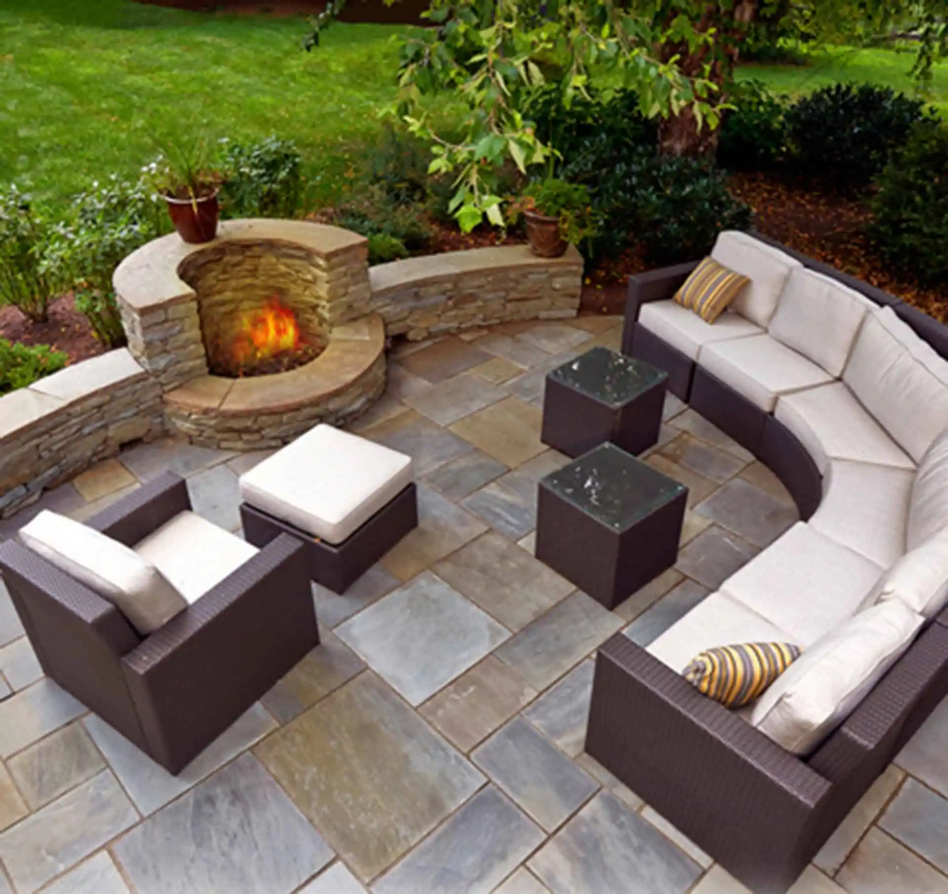 Outdoor patio with a curved stone fire pit, gray sectional sofa with beige cushions, armchair, ottoman, and two small square tables on a tiled patio, surrounded by greenery and trees.
