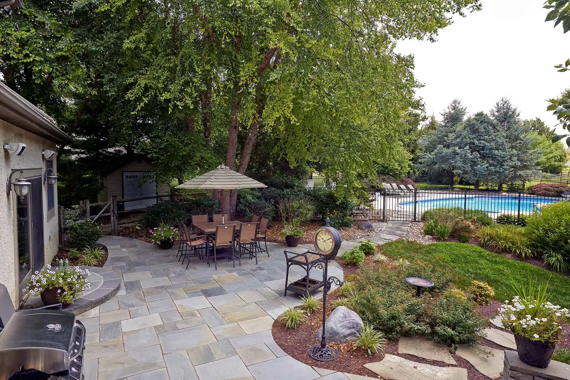 A backyard patio with stone flooring features a dining table and chairs under an umbrella, surrounded by lush greenery. A swimming pool is visible beyond a black fence, with trees and landscaped plants throughout the area.