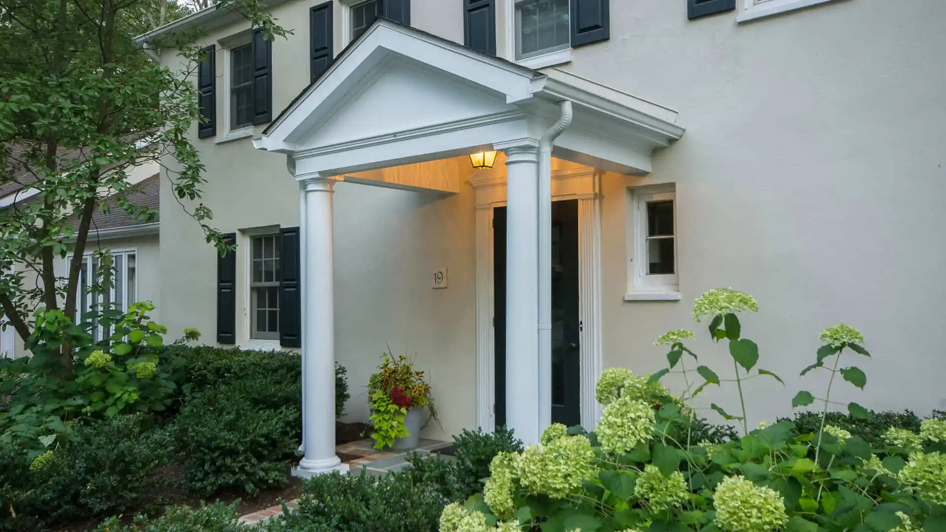 The front entrance of this house features white columns and a triangular pediment, warmly illuminated. Lush green shrubs and blooming hydrangeas enhance the picturesque view, showcasing the exquisite touch of landscaping services in Delaware County, PA.