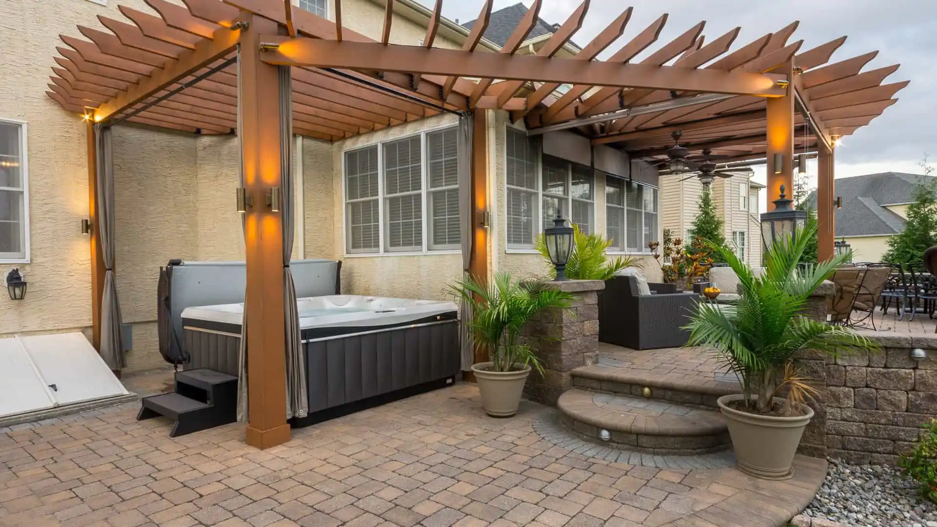 A backyard patio in Delaware County, PA, offers an inviting outdoor living space with a pergola-covered hot tub illuminated by soft lights. Potted plants and wicker furniture enhance the cozy vibe. Steps lead to a seating area, while a dining set completes this beautifully landscaped retreat.
