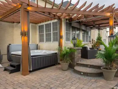 A backyard patio in Delaware County, PA, offers an inviting outdoor living space with a pergola-covered hot tub illuminated by soft lights. Potted plants and wicker furniture enhance the cozy vibe. Steps lead to a seating area, while a dining set completes this beautifully landscaped retreat.