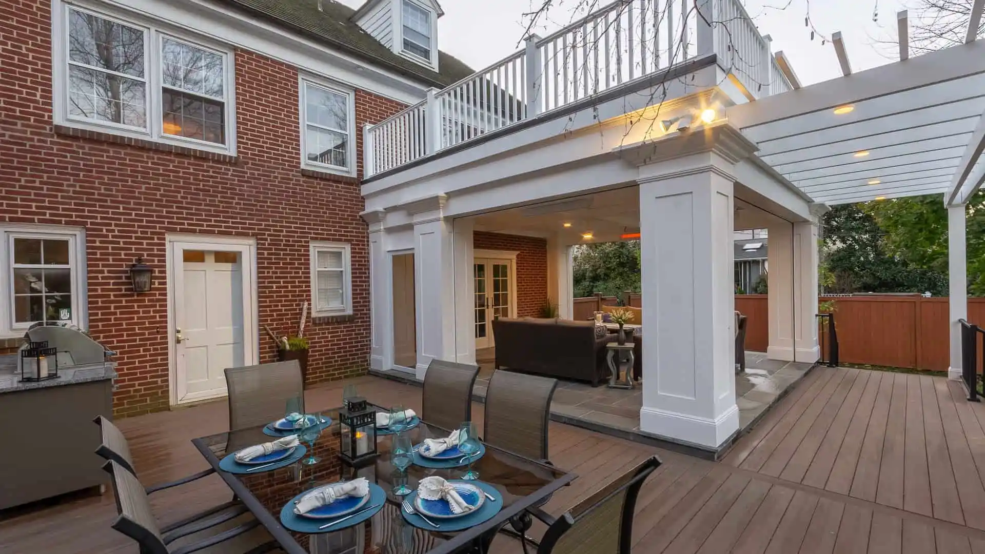 This outdoor living space in Delaware County, PA, boasts a wooden deck dining area for six under a pergola. The setup includes comfortable furniture by a brick house with large windows and a nearby grill. Surrounded by lush landscaping, it’s perfect for relaxation or entertainment.