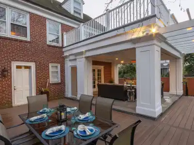 This outdoor living space in Delaware County, PA, boasts a wooden deck dining area for six under a pergola. The setup includes comfortable furniture by a brick house with large windows and a nearby grill. Surrounded by lush landscaping, it’s perfect for relaxation or entertainment.