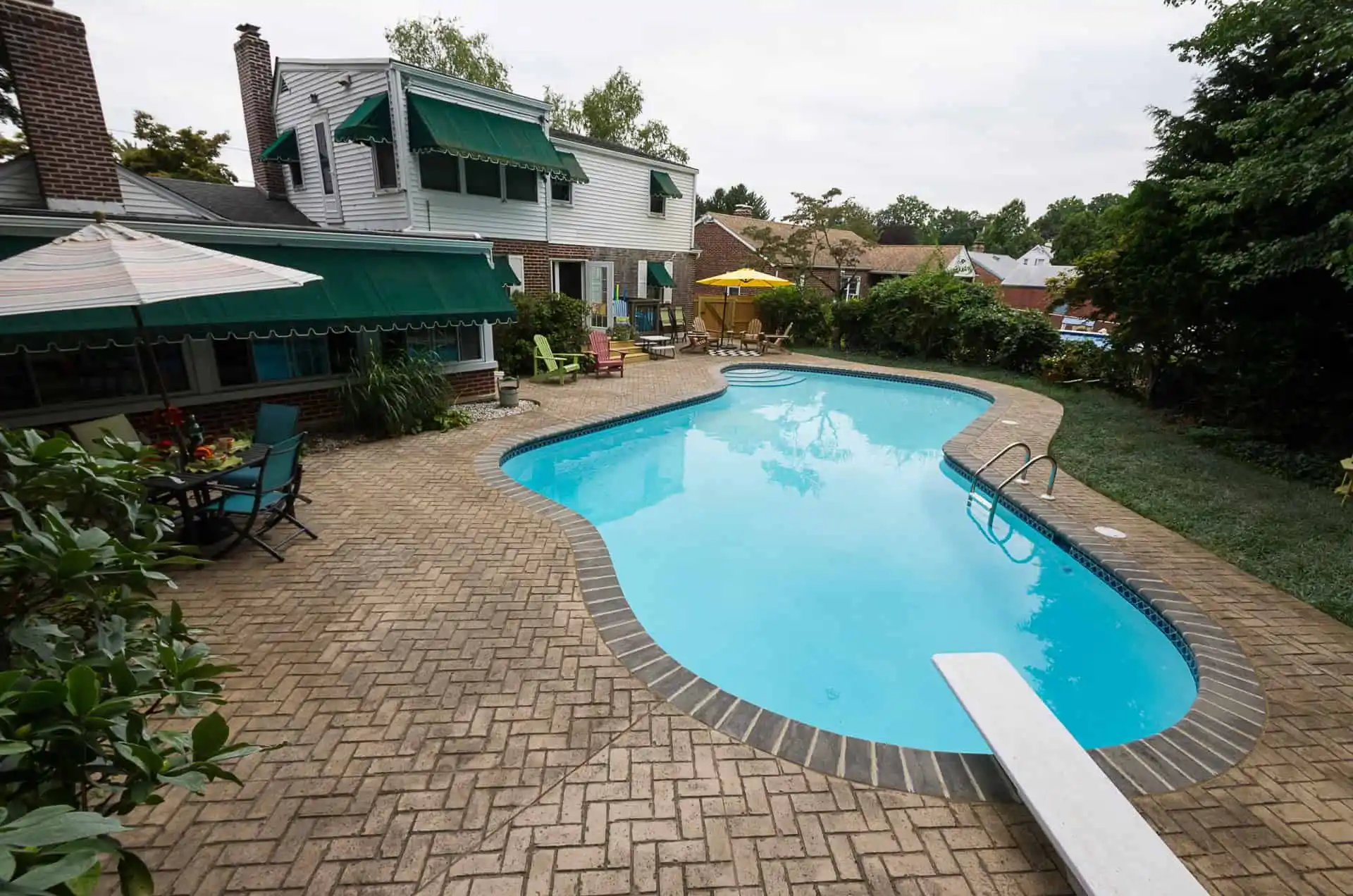 A backyard swimming pool with a diving board, surrounded by brick patio, patio furniture, umbrellas, and greenery. A two-story house with green awnings is in the background under a cloudy sky.