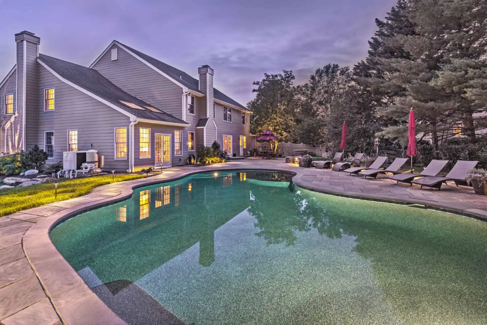 A large backyard in Delaware County, PA, features a pool with gentle lighting. Surrounding the pool are lounge chairs with red umbrellas and lush greenery designed by premier landscaping services. The adjacent two-story house has glowing windows, creating a warm, inviting atmosphere in the evening light.