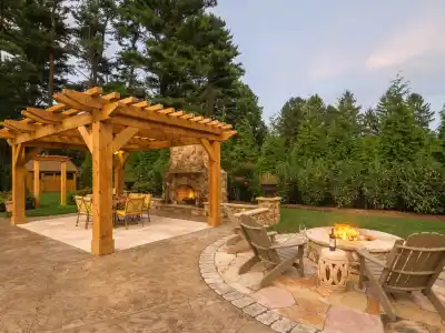 A backyard featuring a wooden pergola over a patio with a dining set. Nearby, a stone fireplace and circular fire pit surrounded by Adirondack chairs create the perfect outdoor living space, crafted with expert hardscaping services in Delaware County, PA, all set against tall trees and hedges.