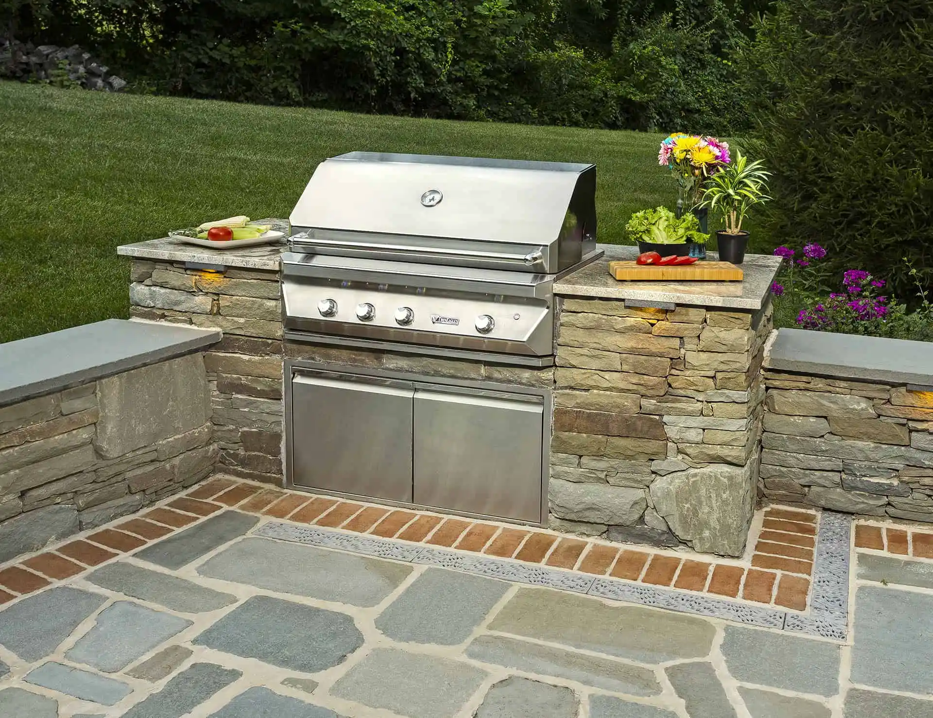 A built-in stainless steel outdoor grill is set in a stone patio with a closed lid. Fresh vegetables and herbs sit on cutting boards to the side, and green grass and trees are in the background.