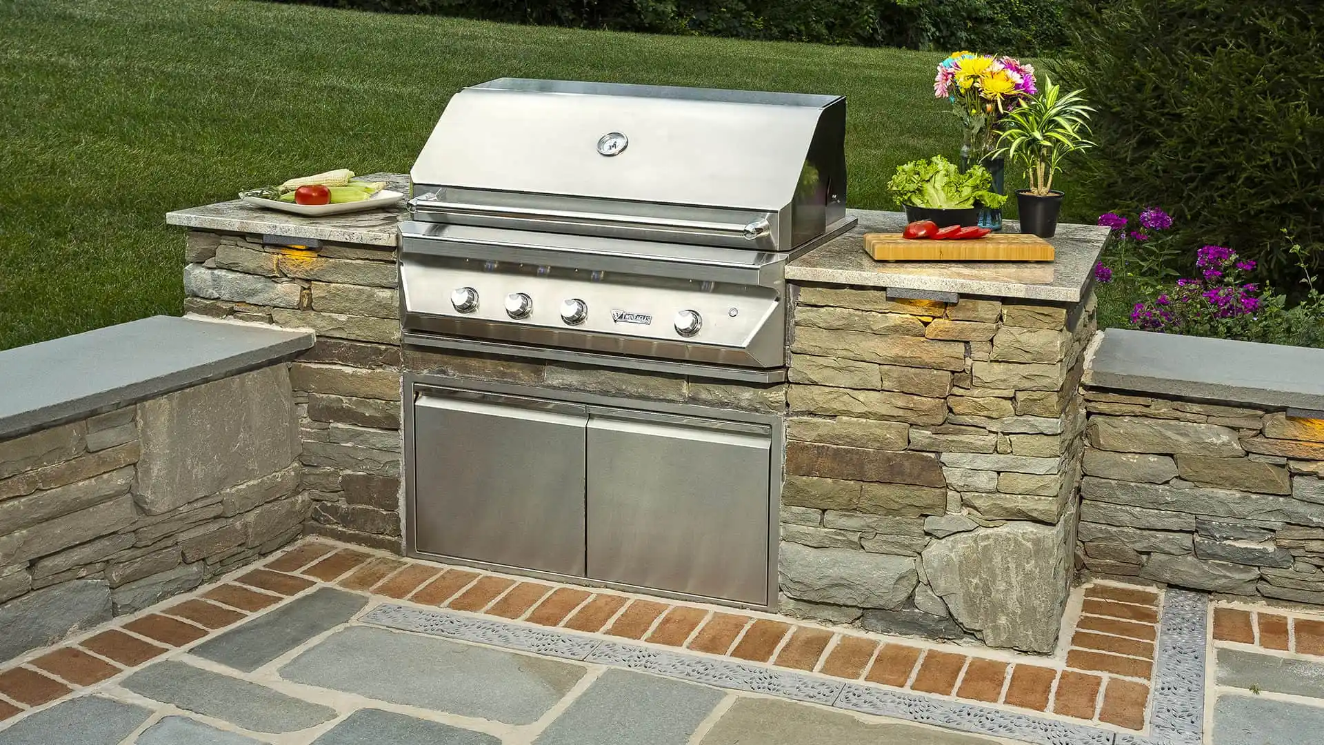 A stainless steel outdoor grill is set within a stone countertop, showcasing expert outdoor living design in Delaware County, PA. It features knobs and a lid, with corn and zucchini sizzling on one side and red peppers on a cutting board nearby. Potted plants and flowers add a natural touch.