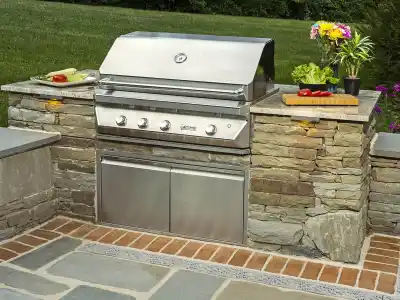 A stainless steel outdoor grill is set within a stone countertop, showcasing expert outdoor living design in Delaware County, PA. It features knobs and a lid, with corn and zucchini sizzling on one side and red peppers on a cutting board nearby. Potted plants and flowers add a natural touch.