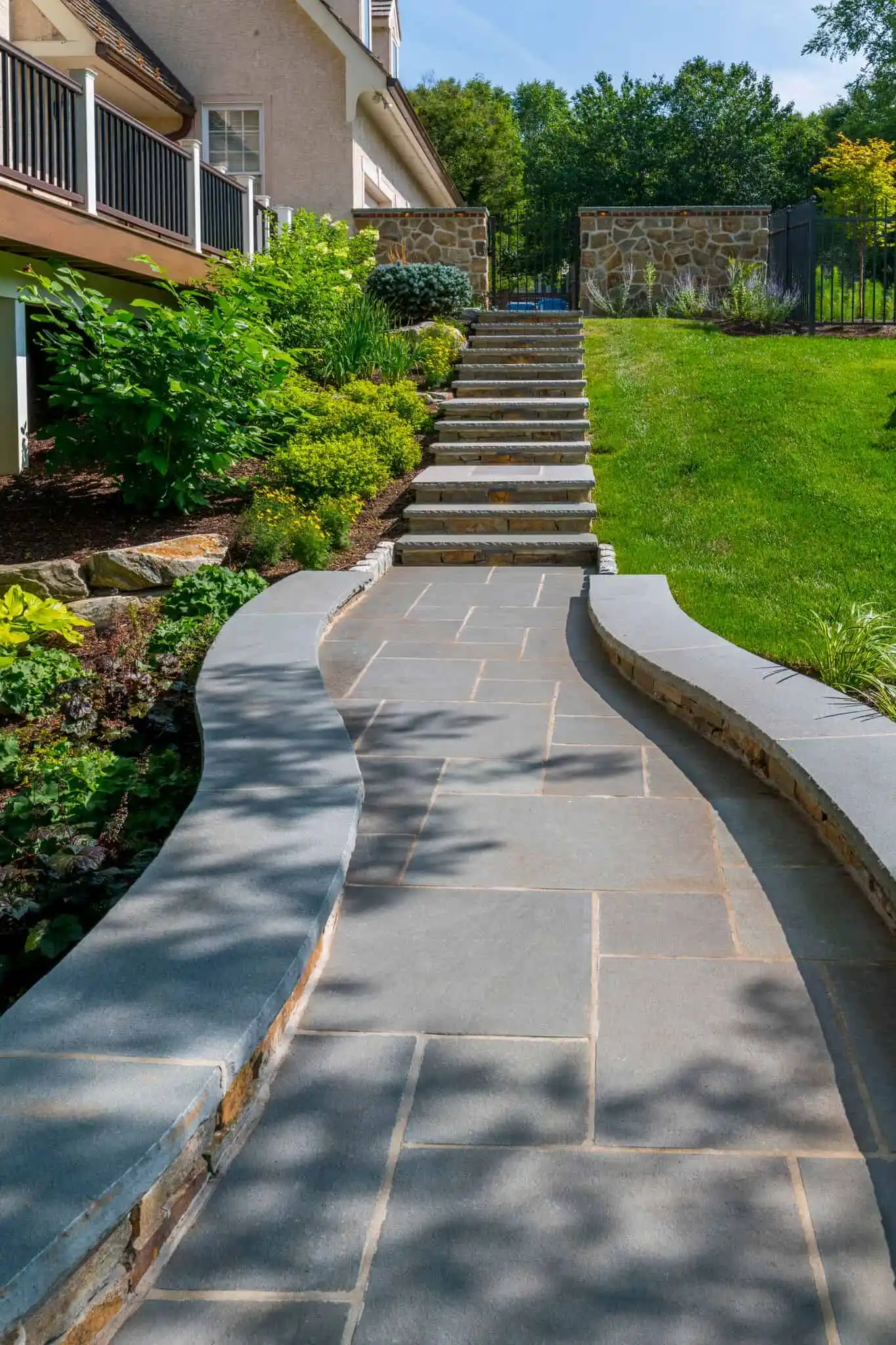 Curved stone pathway with low stone walls, leading to a set of stone steps and a gate, surrounded by green grass, shrubs, and landscaped garden beds.