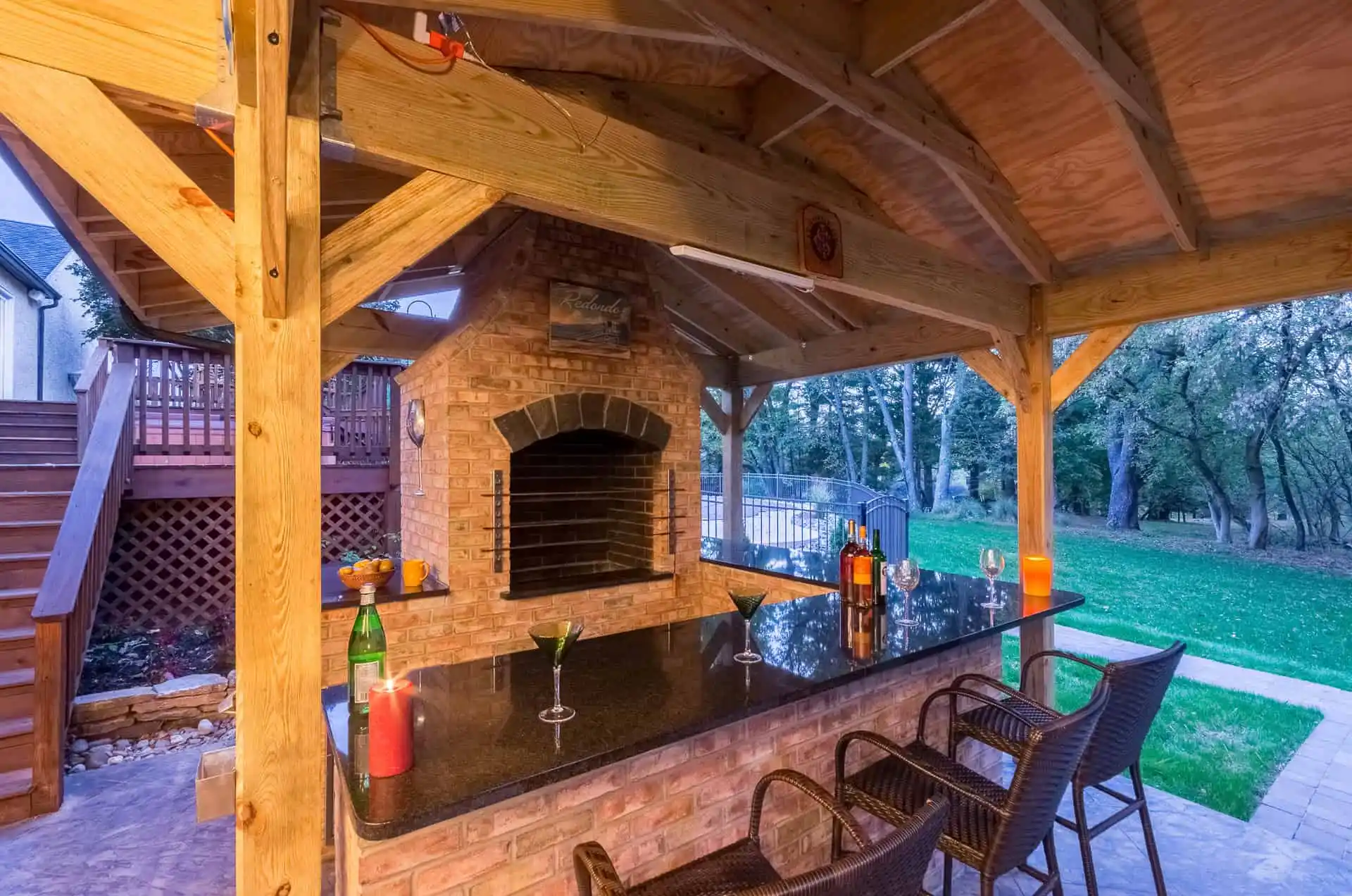 Outdoor bar area under a wooden pergola with brick grill, black countertop, bar stools, drinks, and candles. The space overlooks a grassy yard and is surrounded by trees in the background.