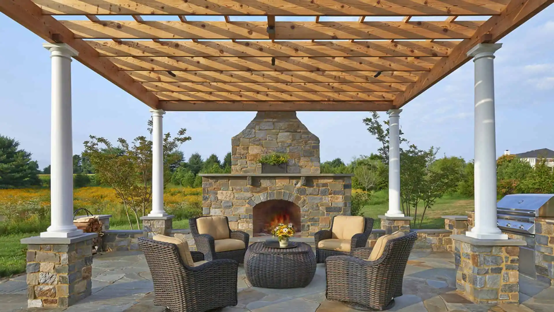 A patio with a wooden pergola, stone fireplace, and four wicker chairs around a round table adorned with flowers offers the perfect outdoor living experience in Delaware County, PA. Surrounded by a grassy field and trees under a clear sky, it showcases excellent landscaping services.