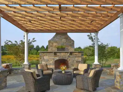 A patio with a wooden pergola, stone fireplace, and four wicker chairs around a round table adorned with flowers offers the perfect outdoor living experience in Delaware County, PA. Surrounded by a grassy field and trees under a clear sky, it showcases excellent landscaping services.