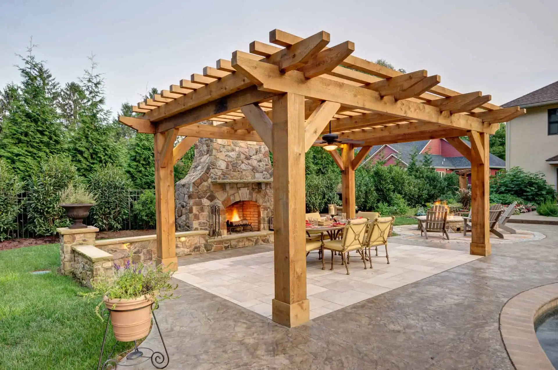A wooden pergola stands over an outdoor dining area with a stone fireplace, surrounded by greenery and a patio with tables, chairs, and potted plants.
