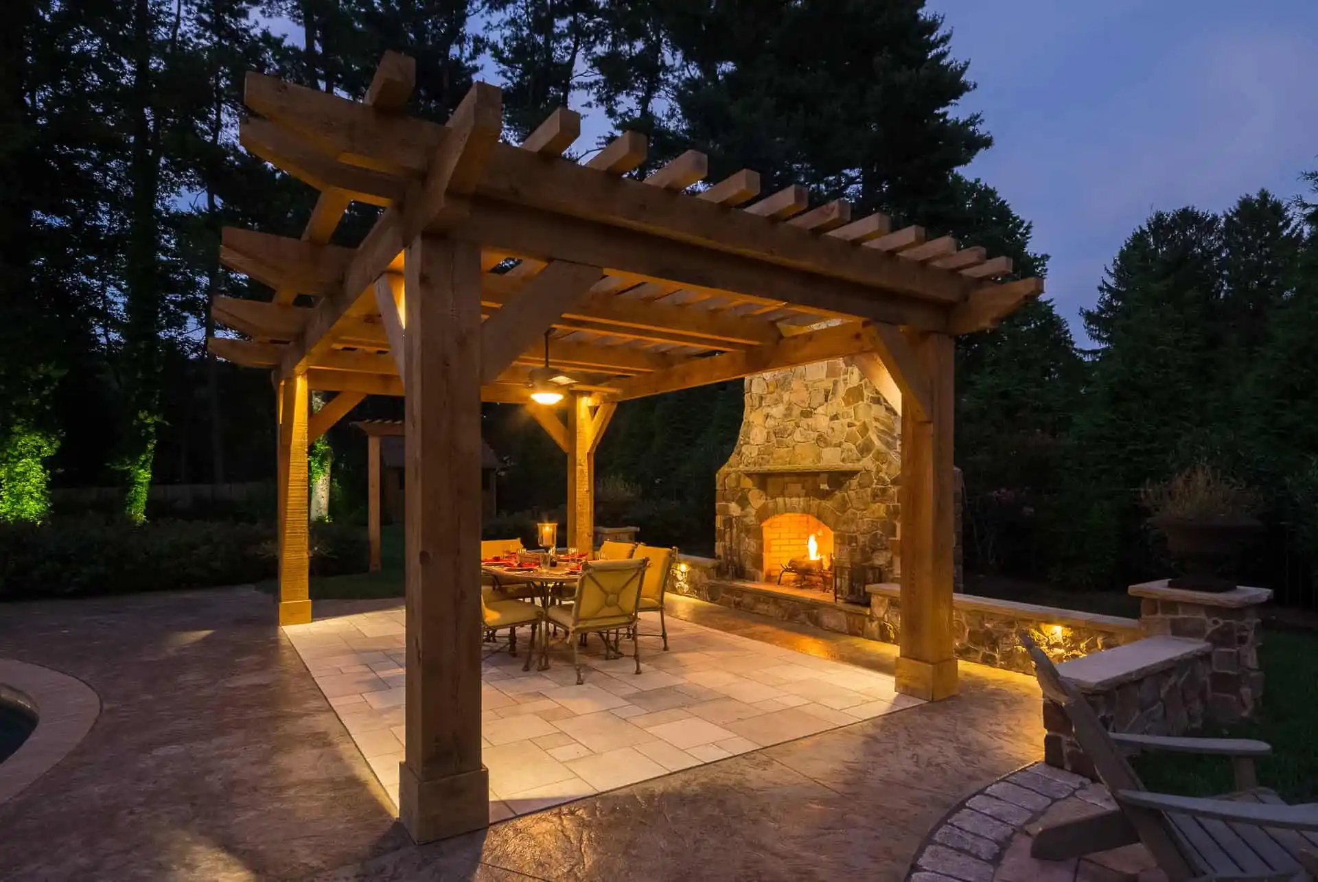 A wooden pergola with a lit stone fireplace, dining table, and chairs sits on a tiled patio at dusk, surrounded by trees and soft landscape lighting for a cozy outdoor atmosphere.