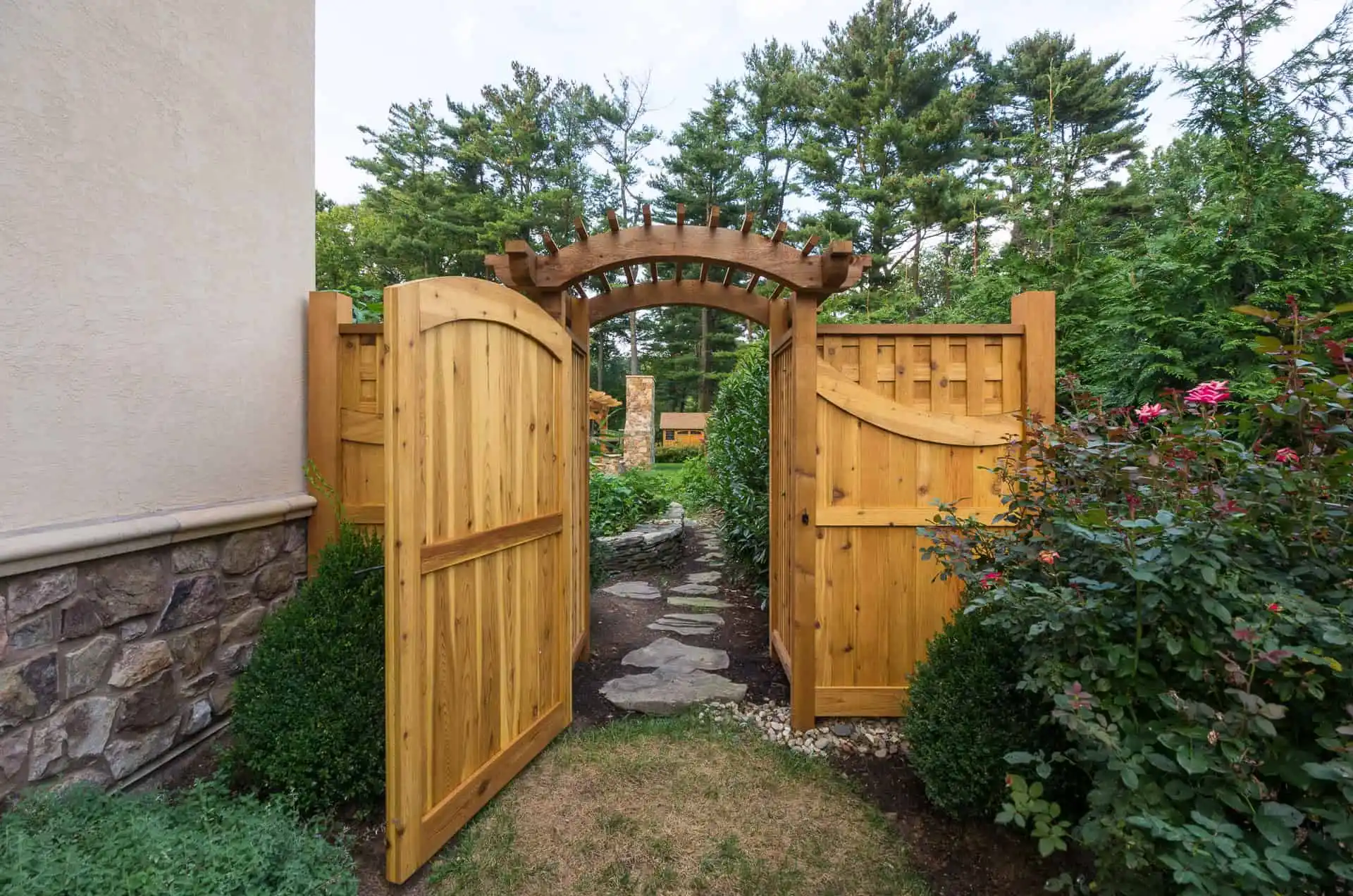 A wooden garden gate with an arched trellis stands open, revealing a stone path bordered by greenery and flowering bushes, leading into a lush, landscaped garden with tall trees in the background.