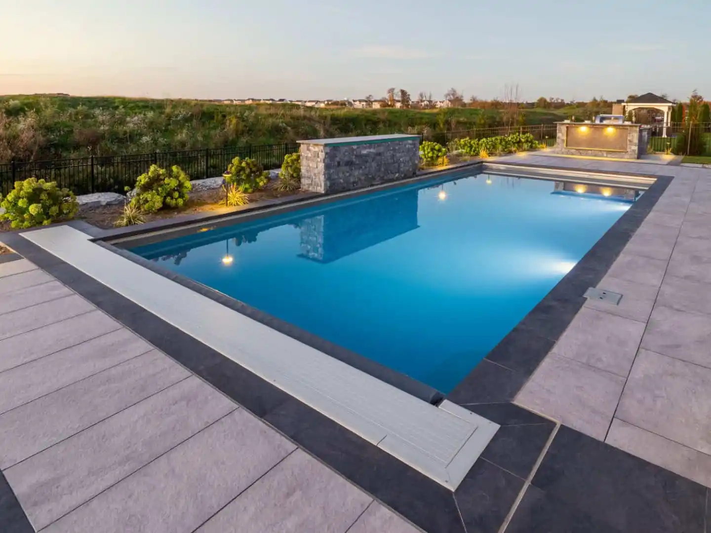 A modern rectangular swimming pool, surrounded by wide gray tiles, reflects the evening sky. In the background, a stone wall with lights enhances the landscaped area featuring greenery and a distant gazebo. Perfect for outdoor living, this Delaware County, PA oasis elevates any hardscape design.