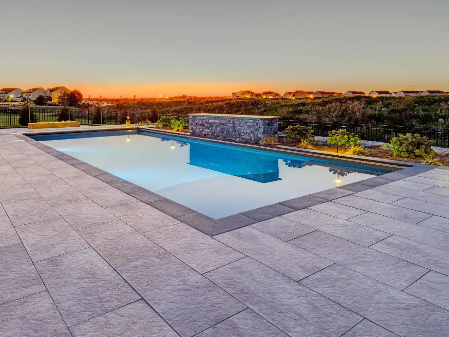 A rectangular swimming pool with clear water reflecting the sunset and a modern water feature at one end. Surrounded by stone tiles from top-notch hardscaping services in Delaware County, PA, it overlooks a grass field and distant houses under a colorful evening sky.