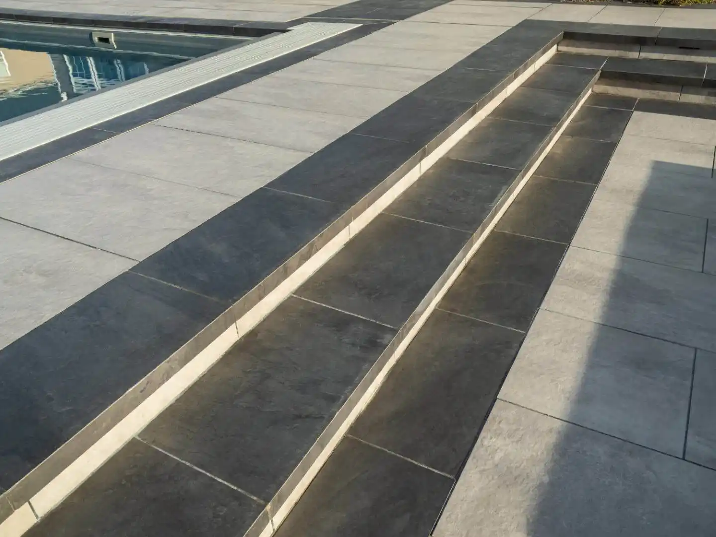 Close-up of a modern outdoor space in Delaware County, PA, featuring light and dark gray stone tiles. The design includes multiple levels with steps leading to an area, possibly a pool, which has a protective cover. Shadows cast across the tiles create a geometric pattern.