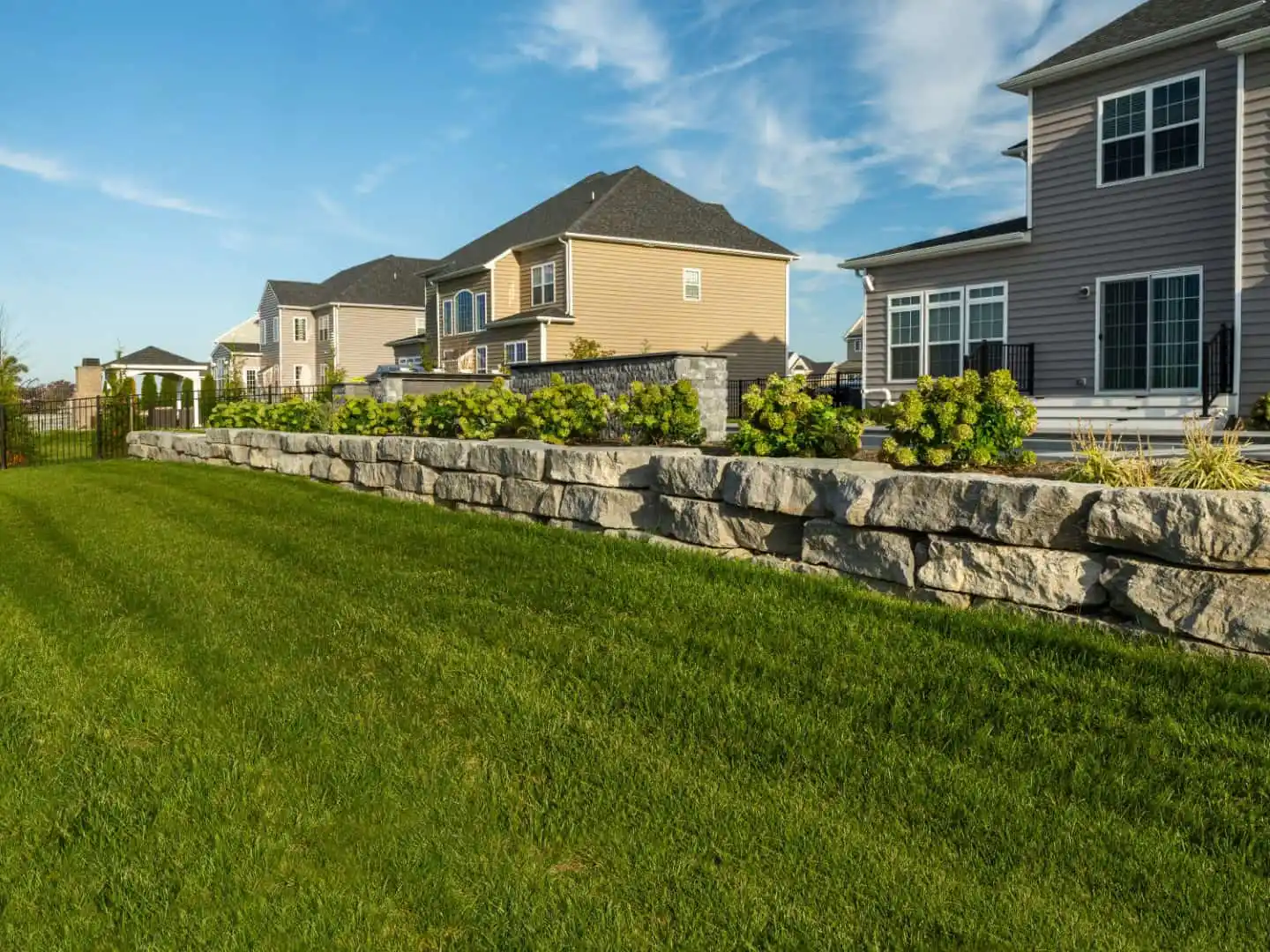 A row of suburban houses with well-maintained lawns is visible. In the foreground, a neatly arranged stone retaining wall supports a garden with green shrubs. The scene showcases expert landscaping services in Delaware County, PA, under a sky that's blue with scattered clouds.