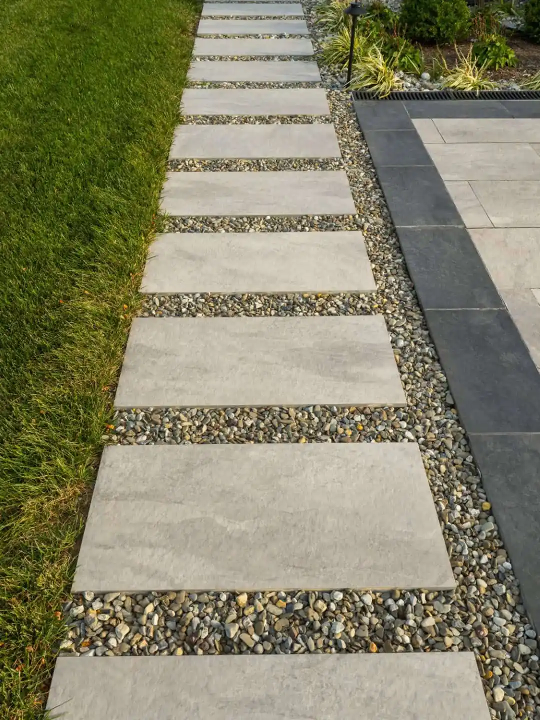 A modern garden pathway with square concrete tiles set in small, gray pebbles. The path is bordered by green grass on one side and a larger paved area on the other, enhanced by decorative plants—ideal for those seeking expert landscaping services in Delaware County, PA.