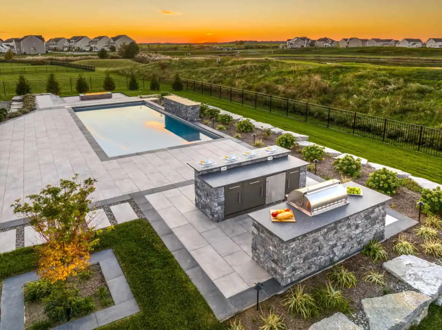 A modern backyard boasts a rectangular pool on a tiled patio. Nearby, an outdoor kitchen with grill and counter space enhances the charm. Expertly crafted by hardscaping services in Delaware County PA, it’s all set under a vibrant orange sunset sky, framed by lush shrubs and grass.