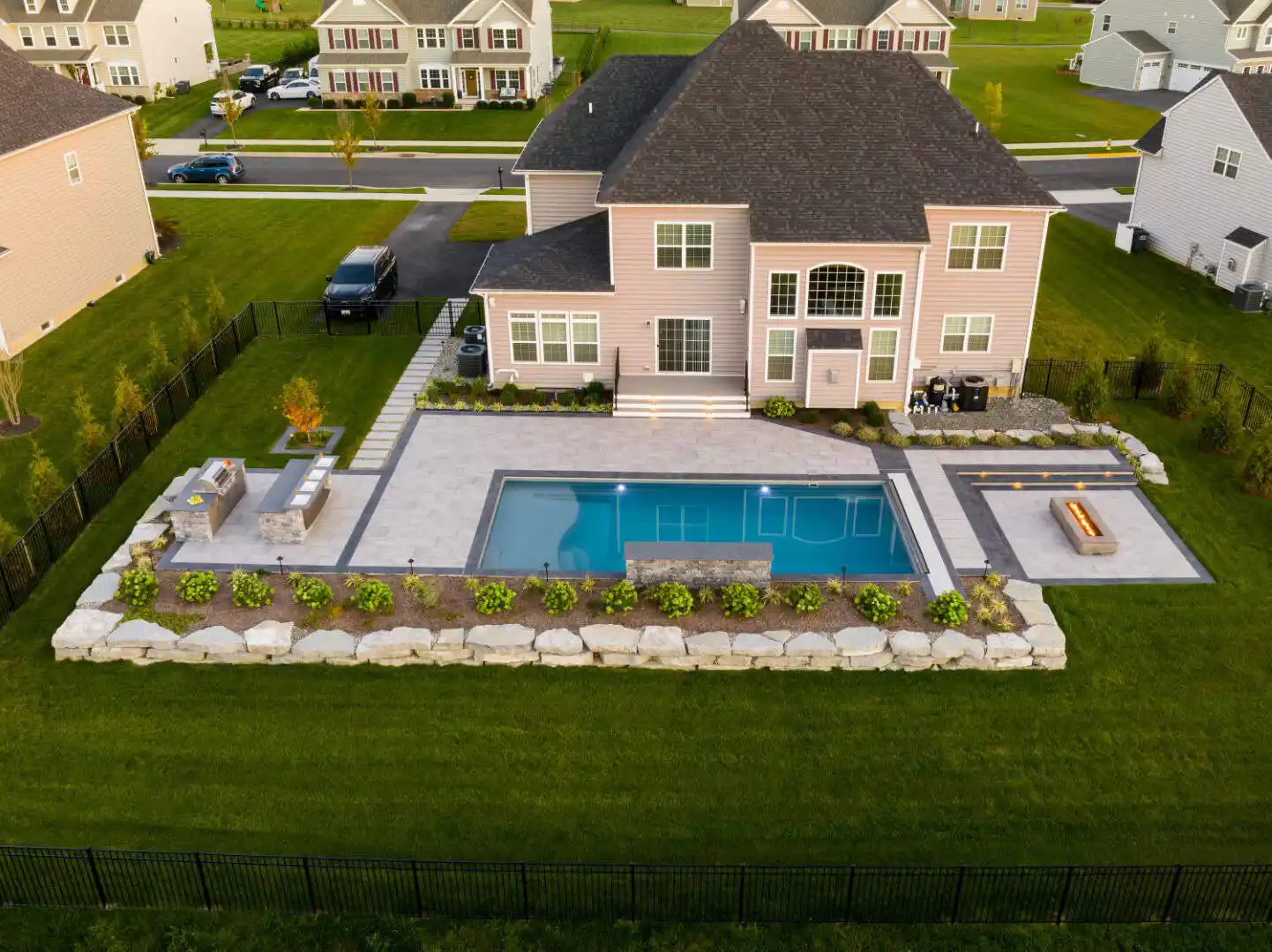 Aerial view of a suburban backyard featuring a modern house with a large pool, showcasing expert landscaping services in Delaware County PA. The yard boasts neatly arranged stone tiles, a fire pit area, landscaped greenery, and outdoor seating. Surrounding houses and road are visible in the background.