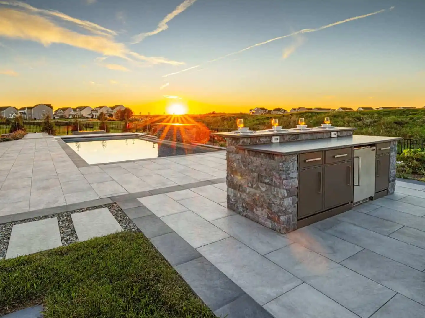 Outdoor patio with a stone kitchen island and stainless steel appliances at sunset. A rectangular pool reflects the sky in this outdoor living haven in Delaware County, PA. The area features light stone tiles bordered by grass, offering hardscaping services with a scenic horizon view.