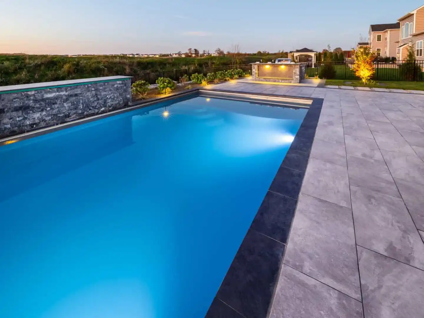 A modern outdoor swimming pool with clear blue water is surrounded by a sleek stone deck, exemplifying top-tier hardscaping services in Delaware County, PA. In the background are a fire pit, a few bushes, and a house under a clear sky, suggesting a serene, early evening setting.A modern outdoor swimming pool with clear blue water is surrounded by a sleek stone deck, exemplifying top-tier hardscaping services in Delaware County, PA. In the background are a fire pit, a few bushes, and a house under a clear sky, suggesting a serene, early evening setting.