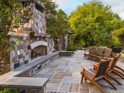 Outdoor living in Delaware County, PA features a patio with a stone fireplace and chimney, surrounded by elegant stone walls. Wooden lounge chairs with cushions face the hearth, while lush greenery creates a serene atmosphere. Perfect for enjoying nature's tranquility.