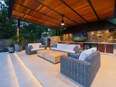 Outdoor patio featuring wicker sofas around a wooden table, with a stone wall backdrop and a TV showcasing the game. Ceiling fans whirl above, and lush greenery thrives. Stairs lead to this cozy outdoor living space in Delaware County, PA, under a pergola adorned with potted plants.