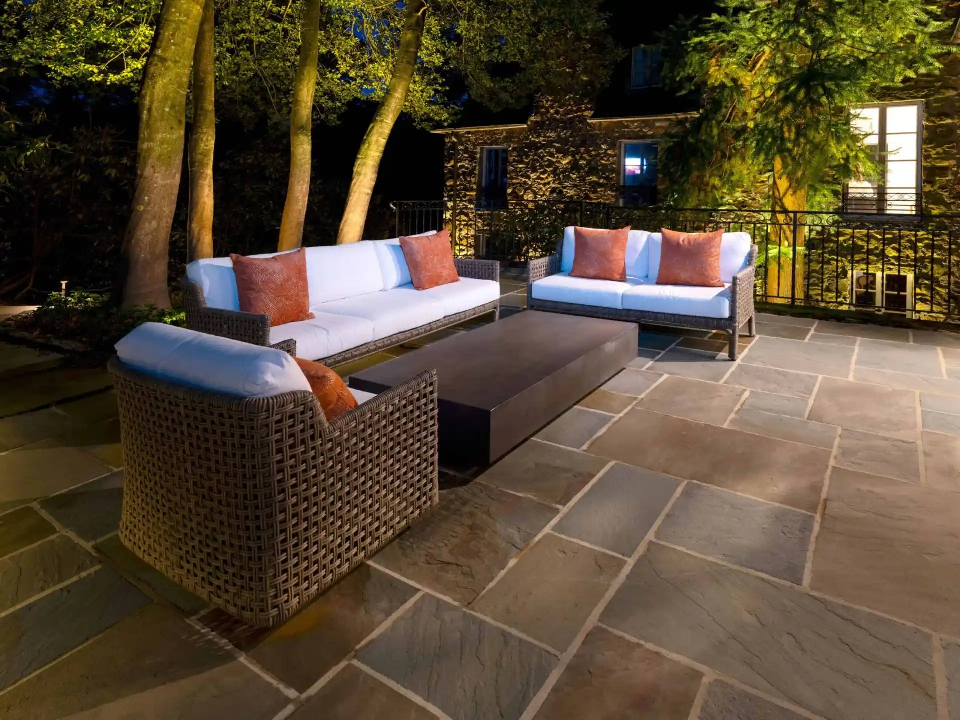 Outdoor living in Delaware County, PA is elevated with this nighttime patio scene. Two wicker sofas and an armchair, adorned with white cushions and brown pillows, encircle a rectangular coffee table on a stone floor. A backdrop of trees and illuminated windows completes the serene setting.
